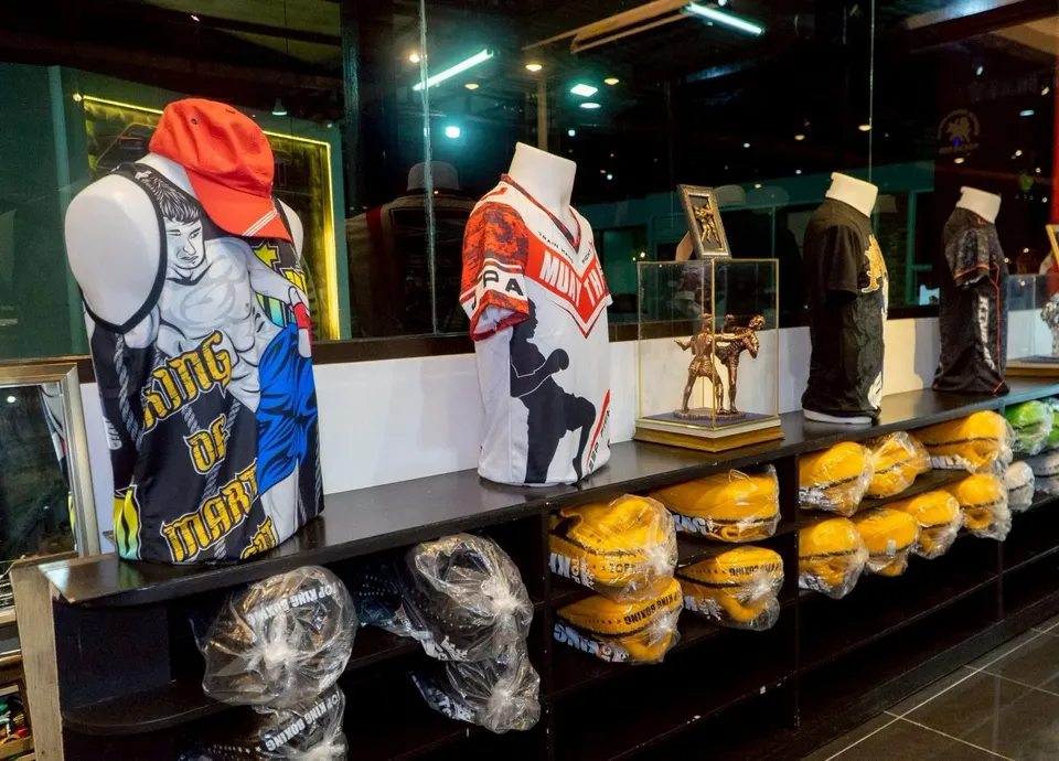 Muay Thai jerseys on mannequins beside a gold fighters statuette in a glass case, with kick pads on the shelf below
