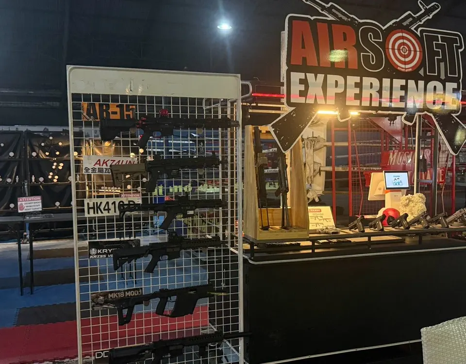 The AIRSOFT EXPERIENCE booth inside Patong Boxing Stadium, with replica rifles on a wire rack and pistols on the counter